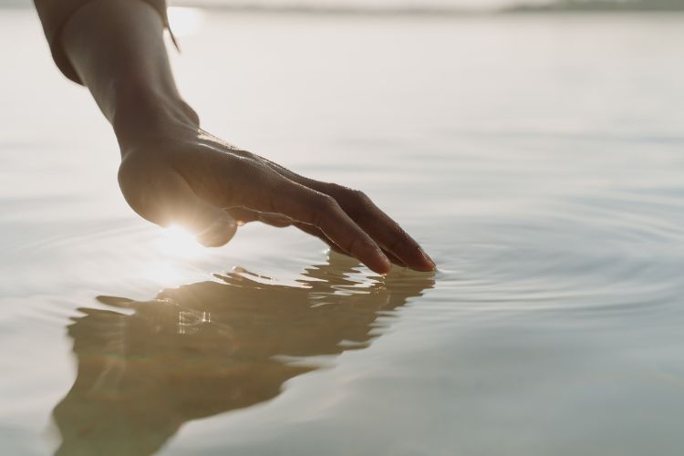 Hand berührt sanft die Wasseroberfläche, Sonnenlicht spiegelt sich im Wasser – Symbol für Präsenz und Achtsamkeit.